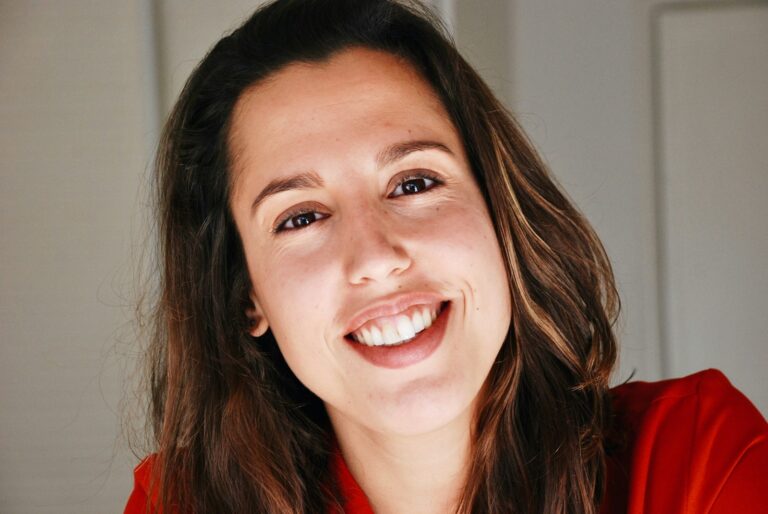 Close-up portrait of a smiling woman with long brown hair wearing a red top, looking at the camera.