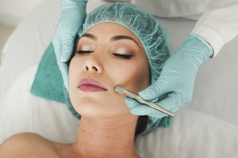 Woman with eyes closed receiving a cosmetic facial treatment; clinician in teal gloves works on her cheek with a metal skin tool while she rests with a disposable cap on his/her head.