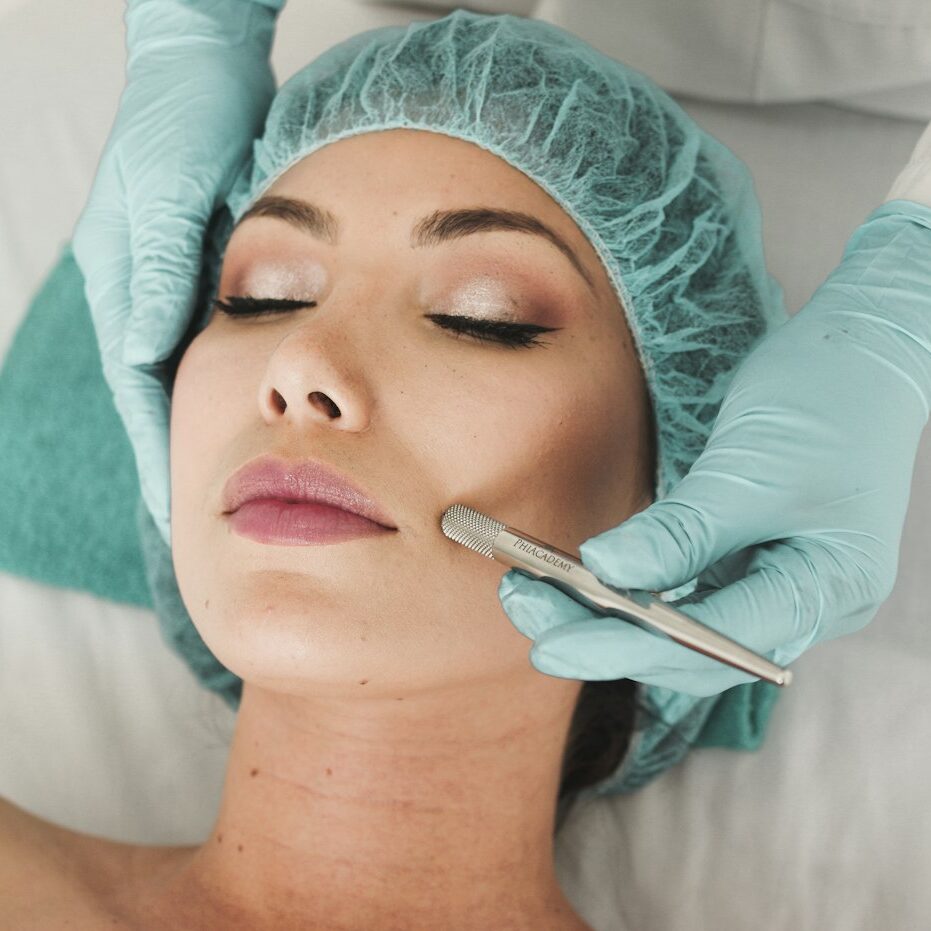 Patient receiving a medical-grade facial peel treatment in Orlando Woman with eyes closed receiving a cosmetic facial treatment; clinician in teal gloves works on her cheek with a metal skin tool while she rests with a disposable cap on his/her head.