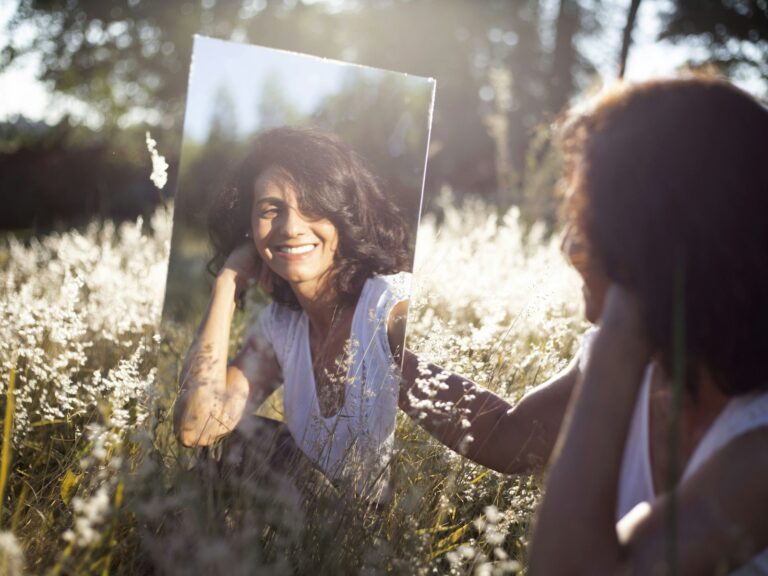 Woman in a white top smiles at her reflection in a handheld mirror in a sunlit meadow with tall grasses